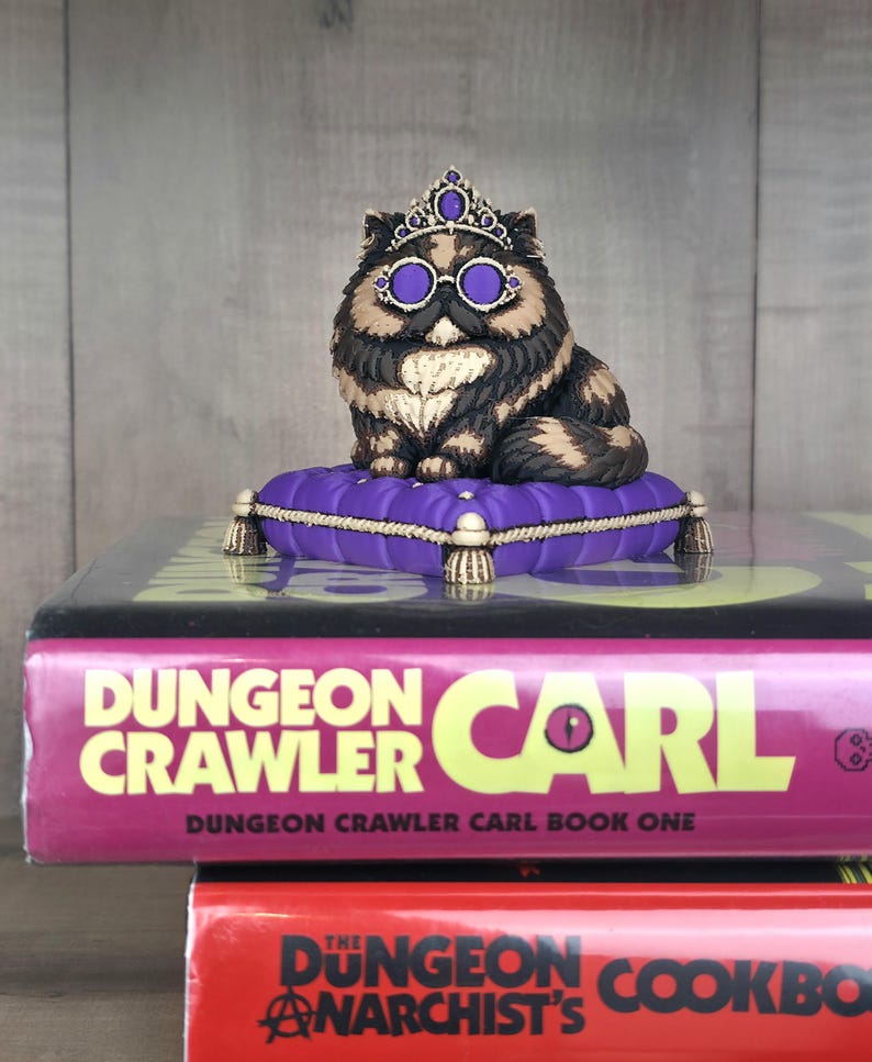 Peut inclure: Figurine d'un chat orn&eacute; d'une couronne et de lunettes, assis sur un coussin violet. Le chat est noir, marron et beige. Le coussin est pos&eacute; sur un livre intitul&eacute; "Dungeon Crawler Carl".