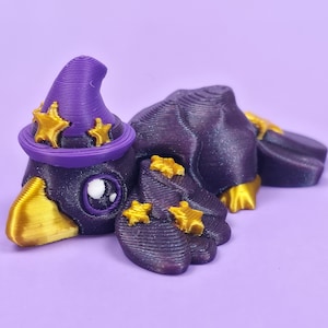 May include: A 3D-printed purple crow figurine wearing a witch hat with gold stars. The crow has gold beak and feet, and a white eye. The figurine is on a purple background.