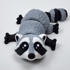 May include: A 3D printed articulated raccoon figurine featuring a gray and white body with black accents around the eyes, nose, and paws. The raccoon has a textured surface, large white-spotted black eyes, and a segmented tail that extends behind.