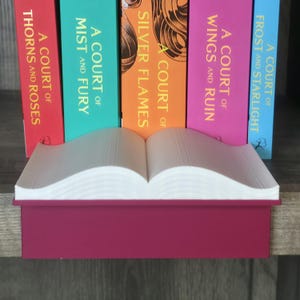 May include: A burgundy-colored book-shaped box with open, white paper pages. Behind it, colorful books with titles like "A Court of Thorns and Roses" and "A Court of Mist and Fury" stand upright, creating a literary display.