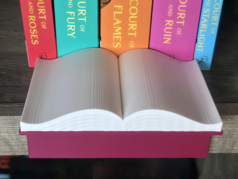 May include: A burgundy-colored book-shaped stand holds an open book with white pages. The stand is positioned in front of a row of colorful books with titles like "Court of Roses" and "Court of Starlight."