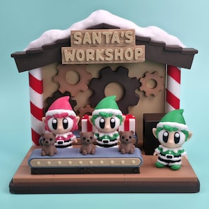 May include: A whimsical Santa's Workshop diorama featuring elf figurines and teddy bears. The scene includes a conveyor belt, gears, and the text "SANTA'S WORKSHOP" on the roof. The elves wear green and red outfits, and the bears are brown.