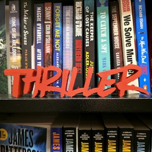 May include: A selection of thriller novels displayed on a shelf, with a bold red "THRILLER" sign. The books feature authors like James Patterson and Richard Osman. The spines are varied in colour, with titles such as "The September House" and "To Catch a Spy".