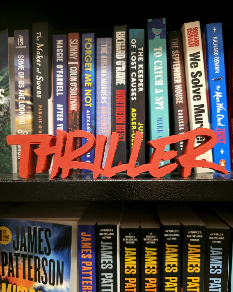 May include: A selection of thriller novels displayed on a shelf, with a bold red "THRILLER" sign. The books feature authors like James Patterson and Richard Osman. The spines are varied in colour, with titles such as "The September House" and "To Catch a Spy".