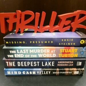 May include: A stack of four thriller novels with a red "THRILLER" sign on top. The book titles are "MISSING, PRESUMED", "THE LAST MURDER AT THE END OF THE WORLD", "THE DEEPEST LAKE", and "HARD CASH VALLEY".