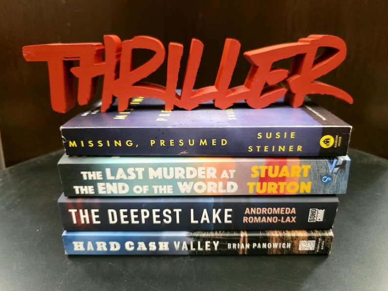 May include: A stack of four thriller novels with a red "THRILLER" sign on top. The book titles are "MISSING, PRESUMED", "THE LAST MURDER AT THE END OF THE WORLD", "THE DEEPEST LAKE", and "HARD CASH VALLEY".