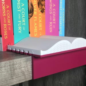 May include: A white and magenta book shelf with a book-shaped design. The shelf is holding several books with colorful spines. The books have titles such as "A Court of Thorns and Roses" and "A Court of Mist and Fury".