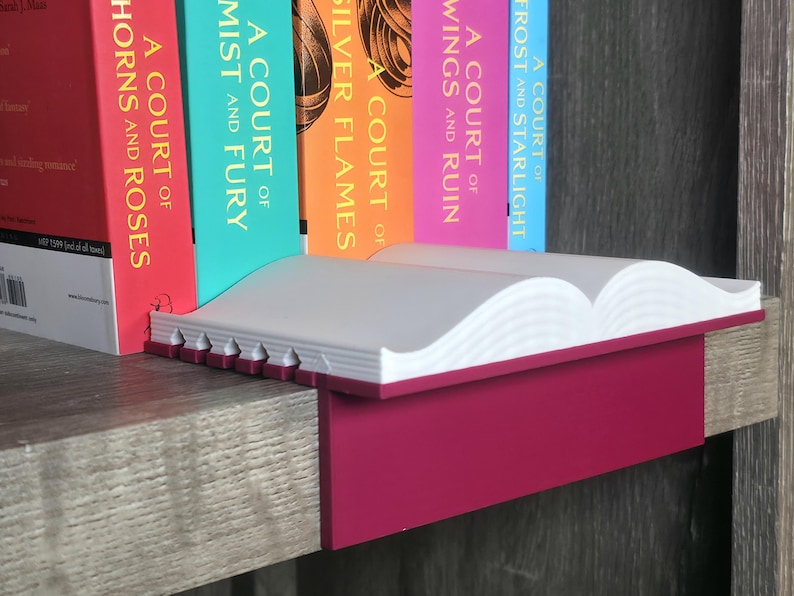 May include: A white and magenta book shelf with a book-shaped design. The shelf is holding several books with colorful spines. The books have titles such as "A Court of Thorns and Roses" and "A Court of Mist and Fury".
