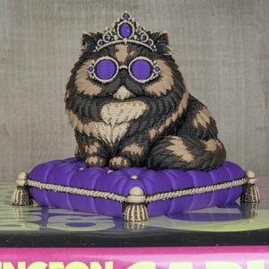 Peut inclure: Figurine d&eacute;corative de chat avec une couronne et des lunettes, assis sur un coussin violet. Le chat a un pelage &agrave; motifs noir, marron et beige. Le coussin a une bordure en corde et des glands, ajoutant une touche d'&eacute;l&eacute;gance.