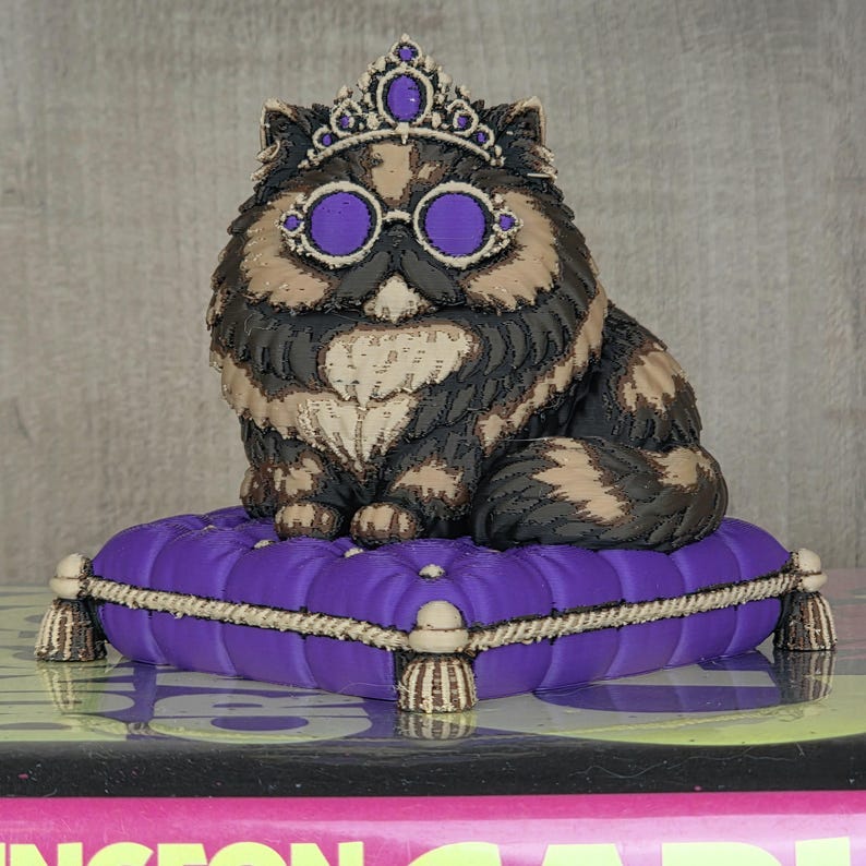 Peut inclure: Figurine d&eacute;corative de chat avec une couronne et des lunettes, assis sur un coussin violet. Le chat a un pelage &agrave; motifs noir, marron et beige. Le coussin a une bordure en corde et des glands, ajoutant une touche d'&eacute;l&eacute;gance.