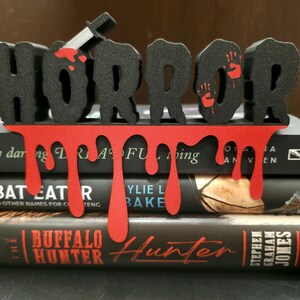 May include: A black "HORROR" sign with a dripping red blood design, a knife, and handprints, placed on top of three books. The books have titles like "The Buffalo Hunter" and "Bat Eat R".