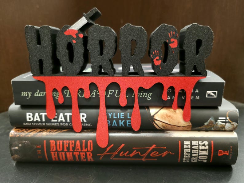 May include: A black "HORROR" sign with a dripping red blood design, a knife, and handprints, placed on top of three books. The books have titles like "The Buffalo Hunter" and "Bat Eat R".