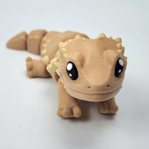 May include: A small, beige, 3D printed figurine of a bearded dragon with large, black eyes. The figurine has a spiky back and is posed in a crawling position.