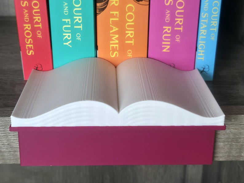 May include: A burgundy-colored book-shaped tray holds a stack of white paper, with the pages open. Behind it, the spines of colorful books are visible, with titles including "Court of Thorns and Roses" and "Court of Fury."