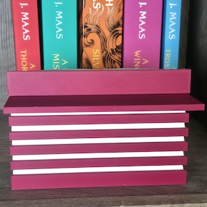 May include: A burgundy-colored book holder with white stripes, holding several books with colorful spines. The books are by Sarah J. Maas, with titles including "A Court of Thorns and Roses" and "A Court of Silver Flames."