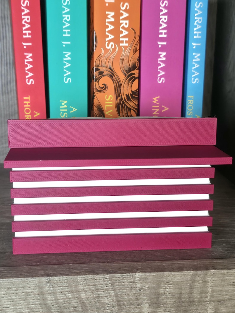 May include: A burgundy-colored book holder with white stripes, holding several books with colorful spines. The books are by Sarah J. Maas, with titles including "A Court of Thorns and Roses" and "A Court of Silver Flames."