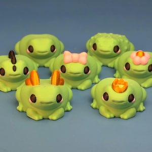 May include: A collection of seven green frog figurines with various decorative elements. Some frogs have orange horns, a pink bow, a flower, or a crown. Each frog has black eyes and is set against a blue background.