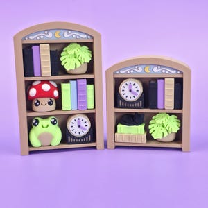 May include: Two miniature bookshelf figurines. One features a red mushroom, a green frog, and a clock. The other has a black cat and a clock. Both have books and a plant. The shelves are tan with a decorative top.