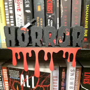 May include: A black and red "HORROR" bookend with a dripping blood design. The letters are black with red accents, including a bloody knife and handprints. The bookend sits in front of a shelf of books.
