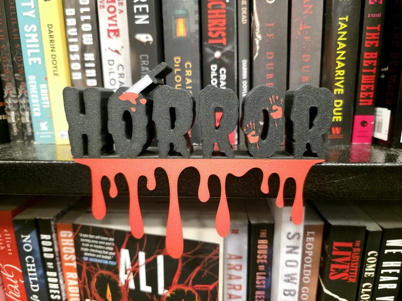 May include: A black and red "HORROR" bookend with a dripping blood design. The letters are black with red accents, including a bloody knife and handprints. The bookend sits in front of a shelf of books.