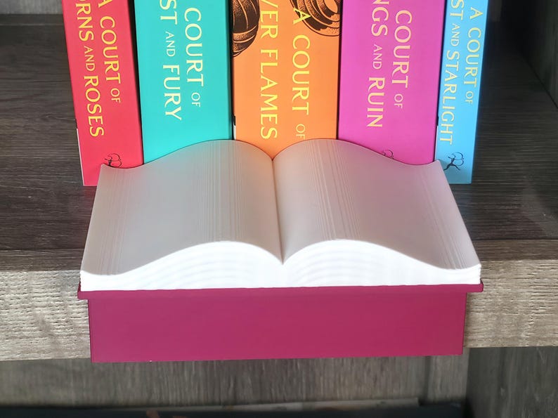 May include: A decorative book-shaped shelf in a deep magenta color, with open pages and a stack of colorful books behind it. The books have titles like "A Court of Thorns and Roses" and "A Court of Mist and Fury".