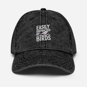 Könnte beinhalten: Eine schwarze Baseballkappe im Vintage-Stil mit dem Text "EASILY DISTRACTED BY BIRDS" in Weiß. In der Mitte ist eine kleine Vogelillustration abgebildet. Die Kappe hat einen gebogenen Schirm und einen Stoffriemenverschluss.