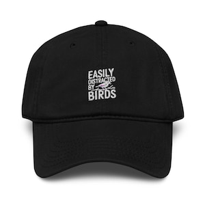 May include: Black baseball cap featuring the embroidered phrase "EASILY DISTRACTED BY BIRDS" in white. The cap has a curved peak and a button on top. A classic, casual hat.