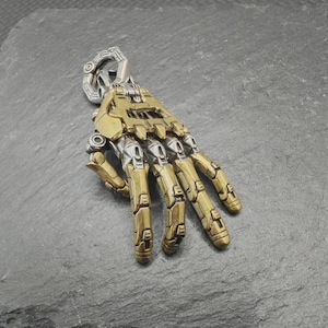 Könnte beinhalten: Anhänger in Form einer mechanischen Hand aus Metall in Gold- und Silbertönen. Die Finger und die Handfläche sind detailliert gestaltet. Die Hand ist leicht geöffnet, mit einer Schlaufe oben zum Aufhängen. Ein originelles und stilvolles Schmuckstück.