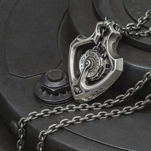 May include: A silver pendant necklace with a detailed, mechanical design. The pendant is shield-shaped with intricate gears and a chain. The necklace is displayed on a dark, textured surface.