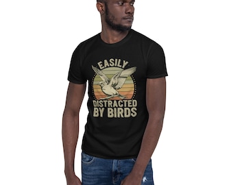 May include: Black t-shirt with a vintage-style graphic featuring a bird in flight against a sunset design. The text reads "EASILY DISTRACTED BY BIRDS" in a distressed font. The shirt is worn by a person.