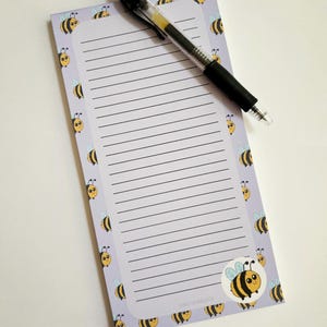 Può includere: Un blocco note viola con un motivo a forma di ape e carta rigata. Una penna nera è appoggiata sul blocco note.