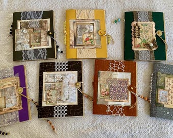 May include: A collection of handmade journals with decorative covers. Each journal features a unique design with vintage-style paper, lace, and embellishments. Colors include green, yellow, purple, brown, and blue. Journals are secured with a button and string closure.