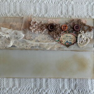May include: A handmade envelope with a vintage aesthetic. It features layers of lace, fabric, and buttons in neutral tones. The envelope is adorned with a decorative label that reads "Savon aux Fleurs de la Rose."