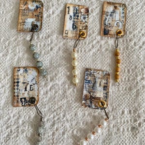 May include: Six vintage-style paper tags with numbers and text, each with a metal ring and a string of beads. The beads are white, gray, and brown.