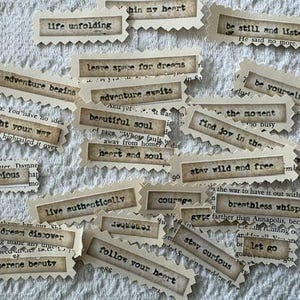 May include: A collection of vintage-style paper tags with inspirational phrases in a typewriter font. Phrases include "life unfolding," "adventure awaits," and "stay wild and free." The tags are arranged on a textured, off-white surface.