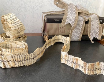 May include: A long strip of book page ribbon, made from old books, is curled and tied in a bow. The ribbon is decorated with a black zig-zag stitch. The ribbon is sitting on a dark surface next to a stack of books wrapped in burlap and lace.