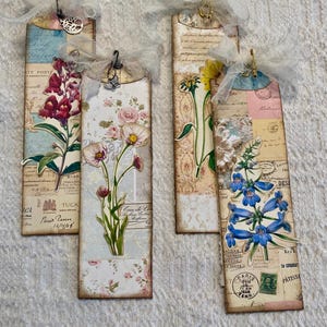 May include: Four handmade bookmarks with floral designs and vintage paper accents. Each bookmark features a different flower, including a pink rose, a blue flower, a yellow flower, and a red flower. The bookmarks are decorated with vintage stamps, paper scraps, and lace.