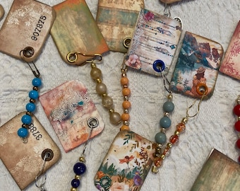 May include: A collection of vintage-style paper tags with various designs and colors, including floral patterns, landscapes, and abstract designs. Each tag has a metal ring and a safety pin attached. The tags are strung together with colorful beads, creating a decorative garland.