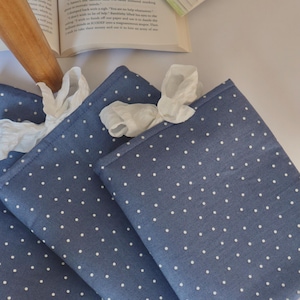 Puede incluir: Cuatro servilletas de tela azules con lunares blancos están extendidas, con servilletas de papel blancas metidas en la parte superior. Las servilletas están sobre una superficie blanca, con un rodillo y un libro abierto en el fondo.