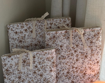 May include: Four book covers with a floral pattern in shades of brown and cream. Each cover is tied with a cream-colored lace ribbon. The covers are arranged in a corner, with a lit candle in the foreground.