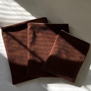 Könnte beinhalten: Drei braune Cordstoffstücke unterschiedlicher Größe liegen auf einer weißen Oberfläche. Der Stoff hat eine gerippte Textur und einen warmen, erdigen Farbton. Die Beleuchtung erzeugt Schatten, die die Textur hervorheben.