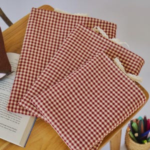 Op de afbeelding: Rood-witte geruite stoffen items met een kanten rand zijn op een houten oppervlak gestapeld. De items lijken op placemats of servetten. Een boek en een mandje met pennen zijn ook zichtbaar.