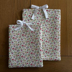 May include: Two rectangular fabric pouches with a floral pattern in pink, yellow, and purple on a cream background. Each pouch is tied with a white ribbon bow. The pouches are of different sizes.