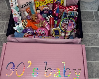 Pode incluir: Uma caixa rosa com as palavras "90's baby" em letras arco-íris. A caixa está aberta e cheia de vários itens com tema dos anos 90, incluindo doces, brinquedos e acessórios.