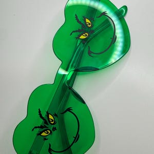 May include: Novelty green sunglasses featuring a Grinch face design. The sunglasses have a translucent green frame and feature the Grinch's face with yellow eyes and a black smile. The lenses are shaped like the Grinch's face.
