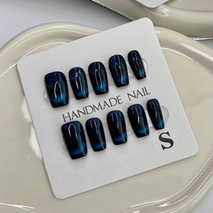 Puede incluir: Un conjunto de diez uñas postizas azul y negro con efecto degradado con el texto "HANDMADE NAIL" y la letra "S" en una tarjeta blanca.