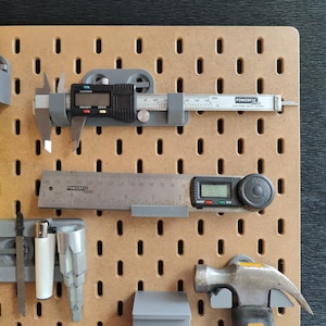 Puede incluir: Un tablero de clavijas con varias herramientas colgadas, incluyendo un calibre digital, una regla, una llave, un martillo y un encendedor. Las herramientas son todas grises y negras.