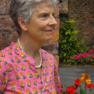 May include: A woman with short grey hair wearing a pink and yellow floral print shirt with a white pearl necklace. She is standing next to a brick wall with a green bush and pink flowers in the background.