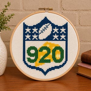 May include: A circular wooden embroidery hoop containing a cross-stitch design of a blue shield shape with white stars, a white football, and the green numbers 920 layered over a yellow cheese wedge shape on a white fabric background.