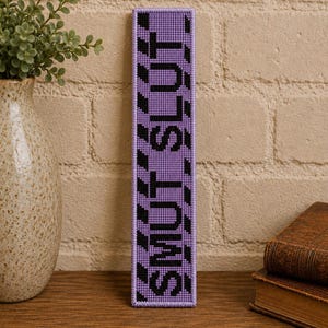 Peut inclure: Un marque-page rectangulaire en point de croix, haut et étroit, avec un fond lavande et une bordure à rayures diagonales noires. Le mot "SMUT" est brodé en lettres capitales noires et grasses verticalement au centre.